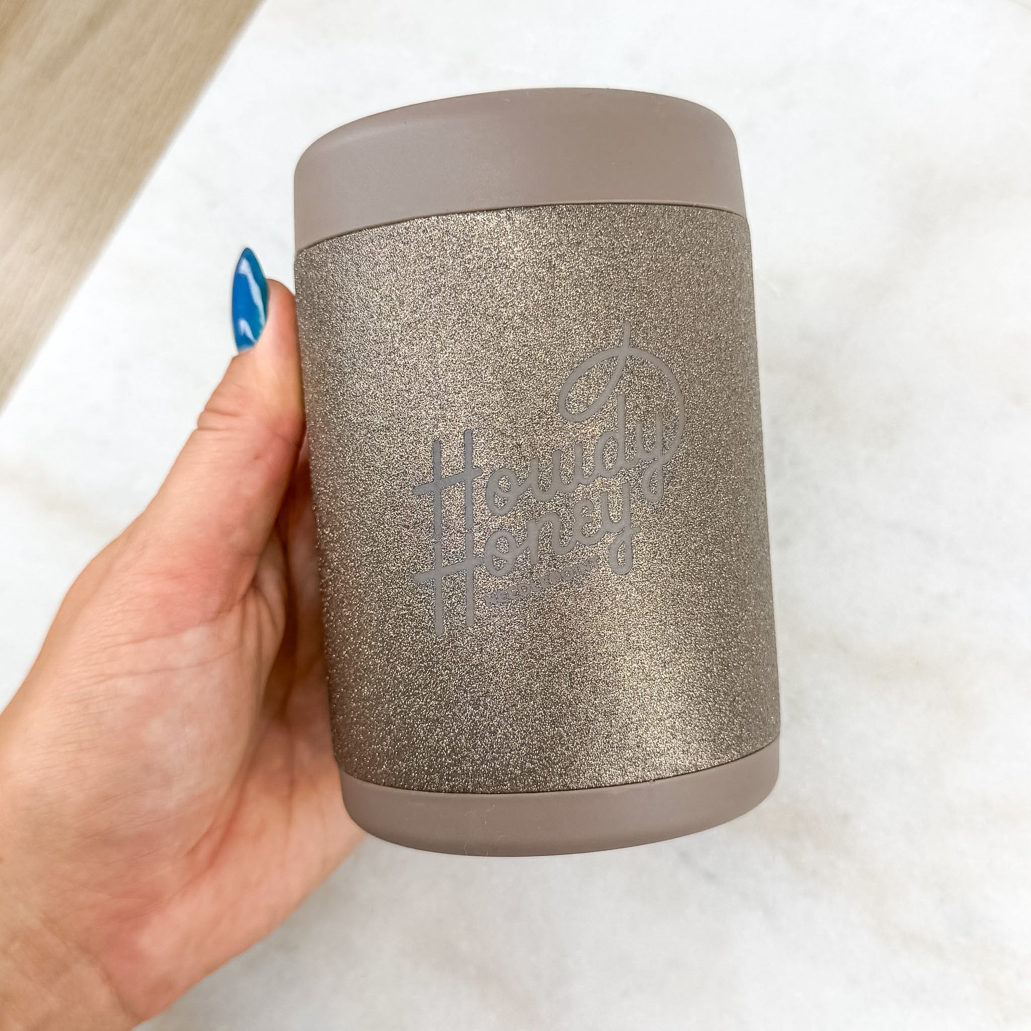 A hand with blue nail polish holds a glittery taupe insulated tumbler with a matching lid. The tumbler features a debossed logo that reads 'The Daily Honey' on the front, set against a light marble background.