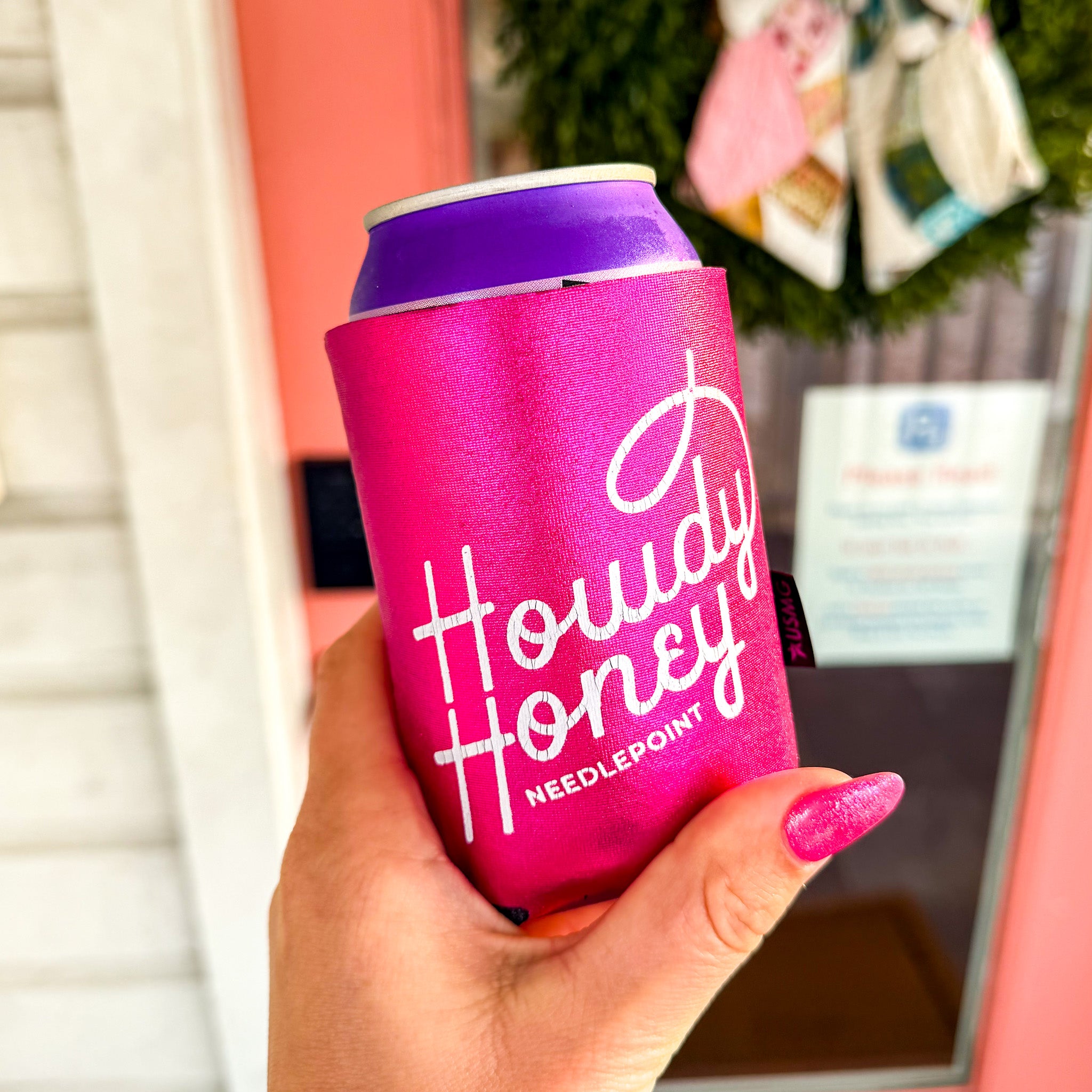 Pink can cooler with 'Howdy Honey' text held by a hand, blurred indoor background
