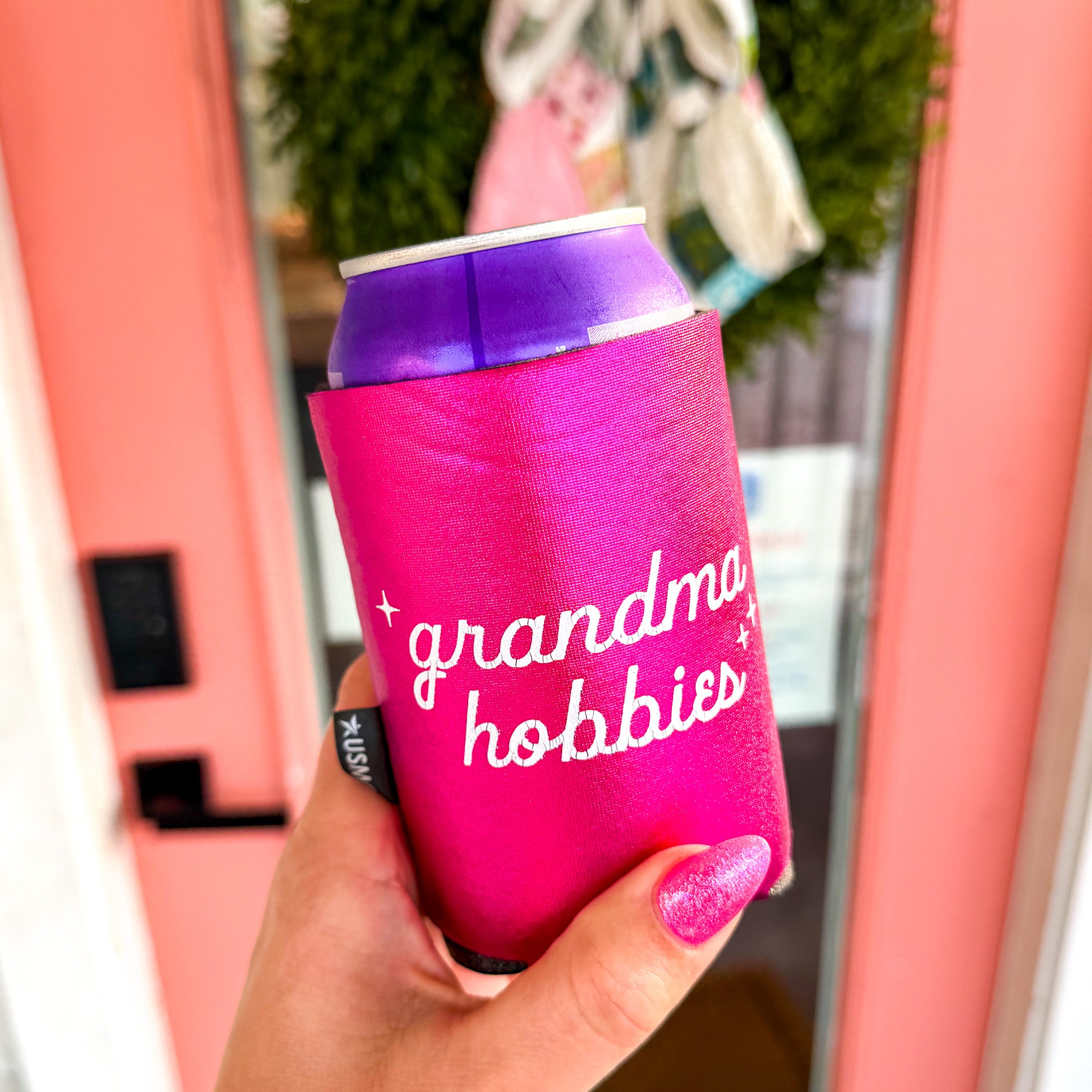 Pink can cooler with 'grandma hobbies' text held by a hand.