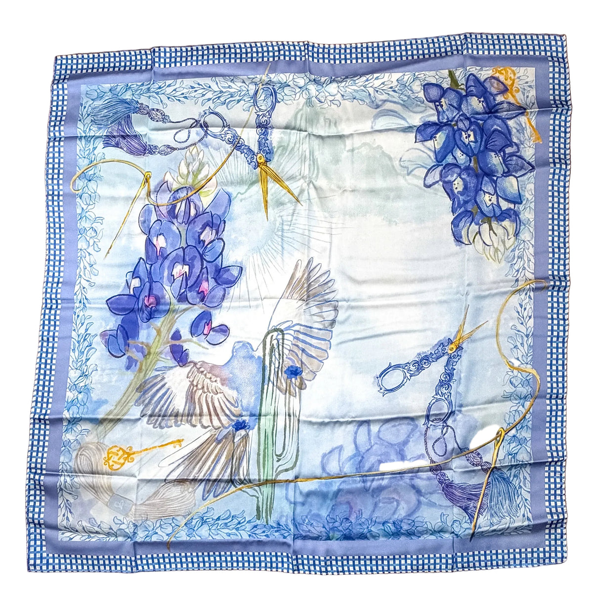 A square silk scarf with a blue and white gingham border, featuring a botanical print of blue lupines, a white bird, and gold abstract line details, laid flat on a wooden surface.