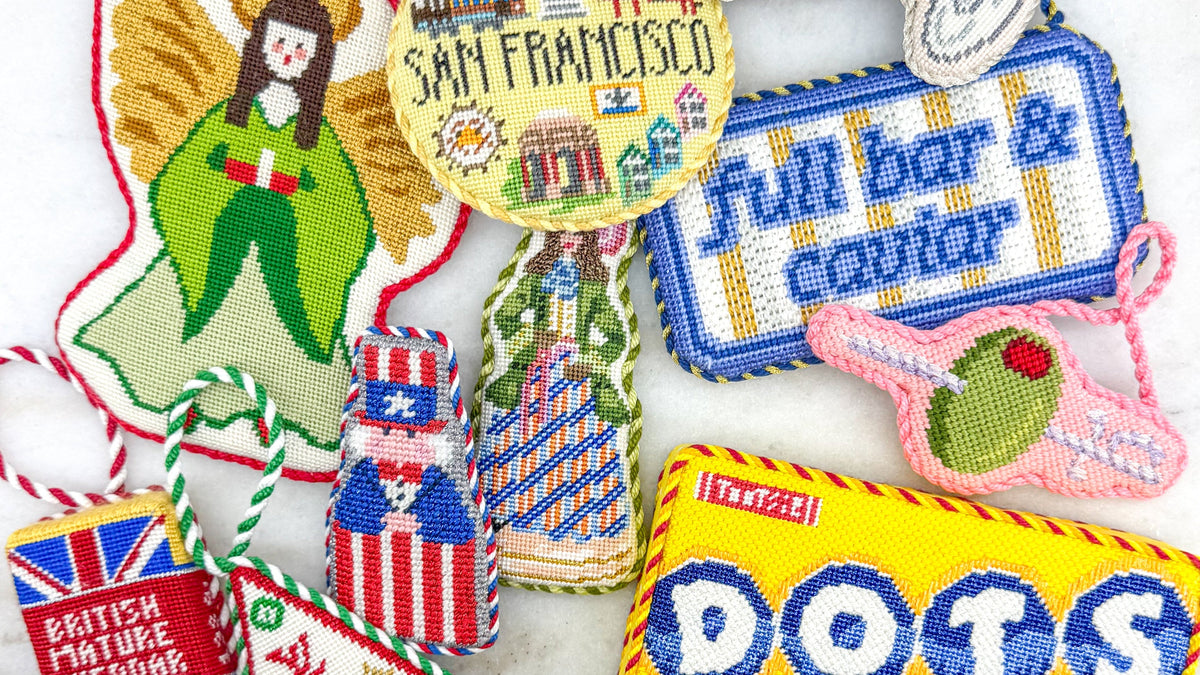 A group of colorful embroidered cross-stitch ornaments and small decorative patches arranged on a white background. The designs include a green angel, a San Francisco-themed round patch, a martini glass, a blue 'Full bar & caviar' sign, a pink oliver, a yellow 'DOTS' patch, a red 'British Mature Cheddar' patch, and several Louis Vuitton monogram patterned pieces.