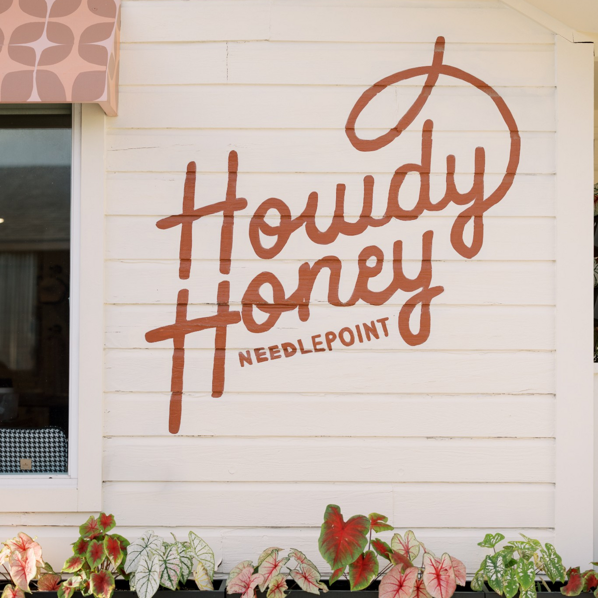 A white exterior wall of a building with large brown script lettering reading 'Howdy Honey' and smaller text 'NEEDLEPOINT' beneath it. A window with a patterned valance is visible on the left, and black planters with pink and green foliage line the bottom of the wall.