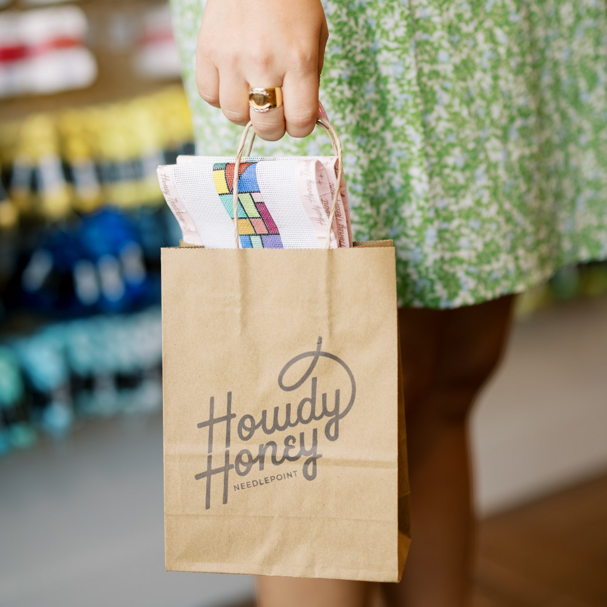 A person in a green floral dress holds a brown paper shopping bag printed with the brand name 'Howdy Honey' in a boutique setting.