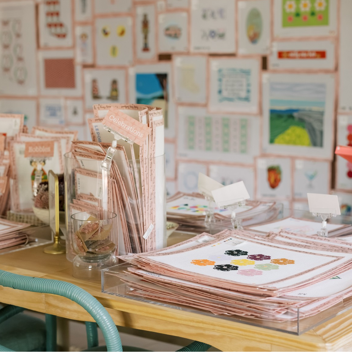 A wooden display table in a craft studio filled with pink and white cardstock sheets, clear plastic holders, and small cards. A teal chair is visible in the foreground, and a wall covered with colorful craft samples and posters is in the background.