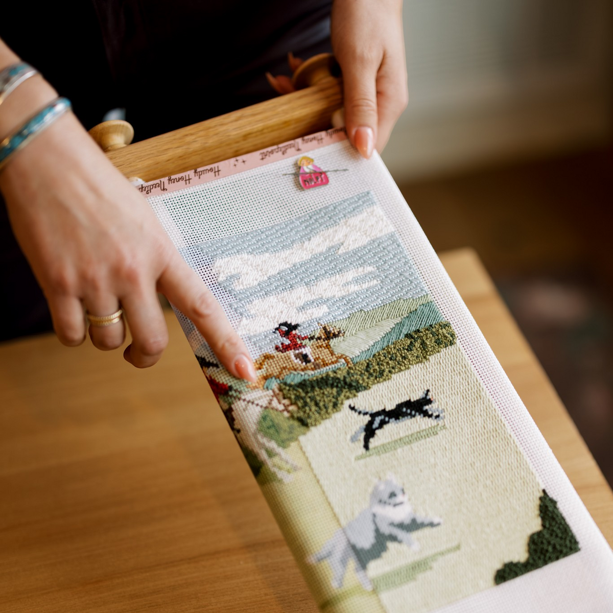 A close-up of a person holding a wooden needle loom with a partially finished cross-stitch piece depicting dogs running through a landscape. The person's hands and part of their torso are visible, and the scene is set on a wooden table.
