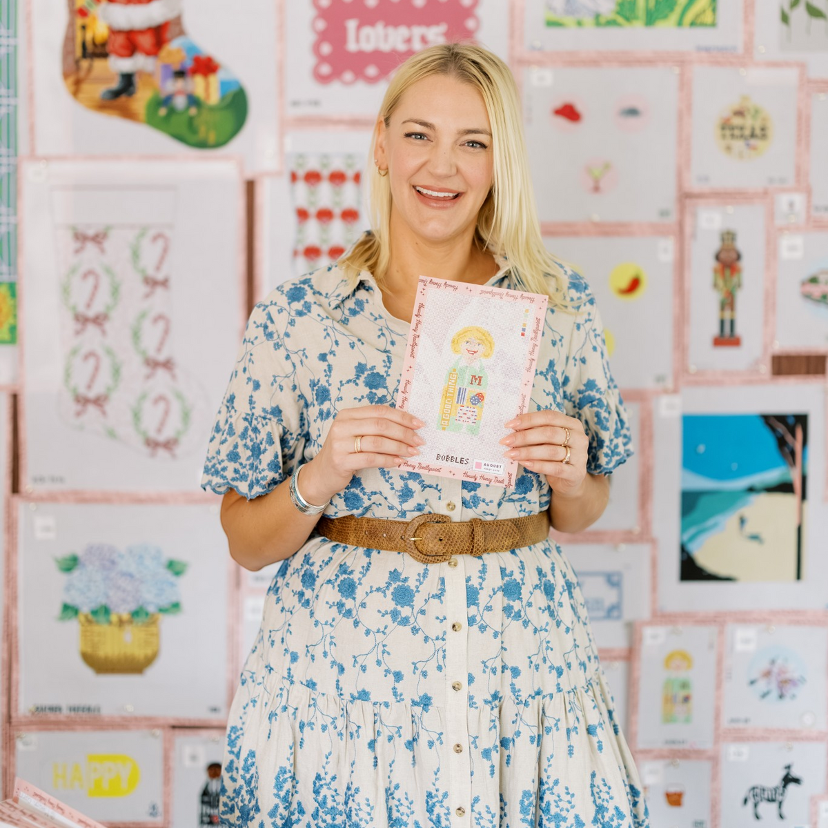 A smiling blonde woman stands in front of a wall covered in colorful greeting cards, holding up a pink and white illustrated card. She is wearing a blue and white floral short-sleeve button-up dress with a brown belt.