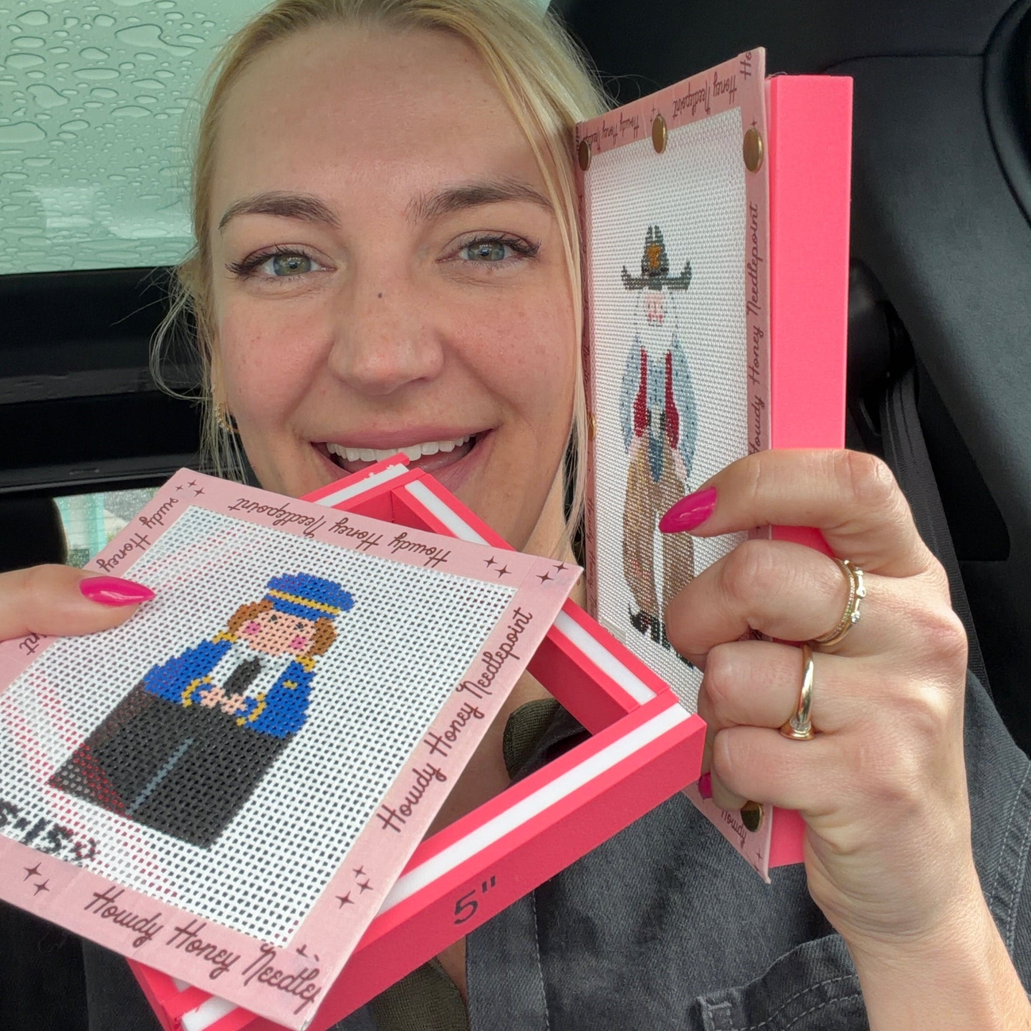 Person holding cross-stitch kits with a smile, inside a vehicle.