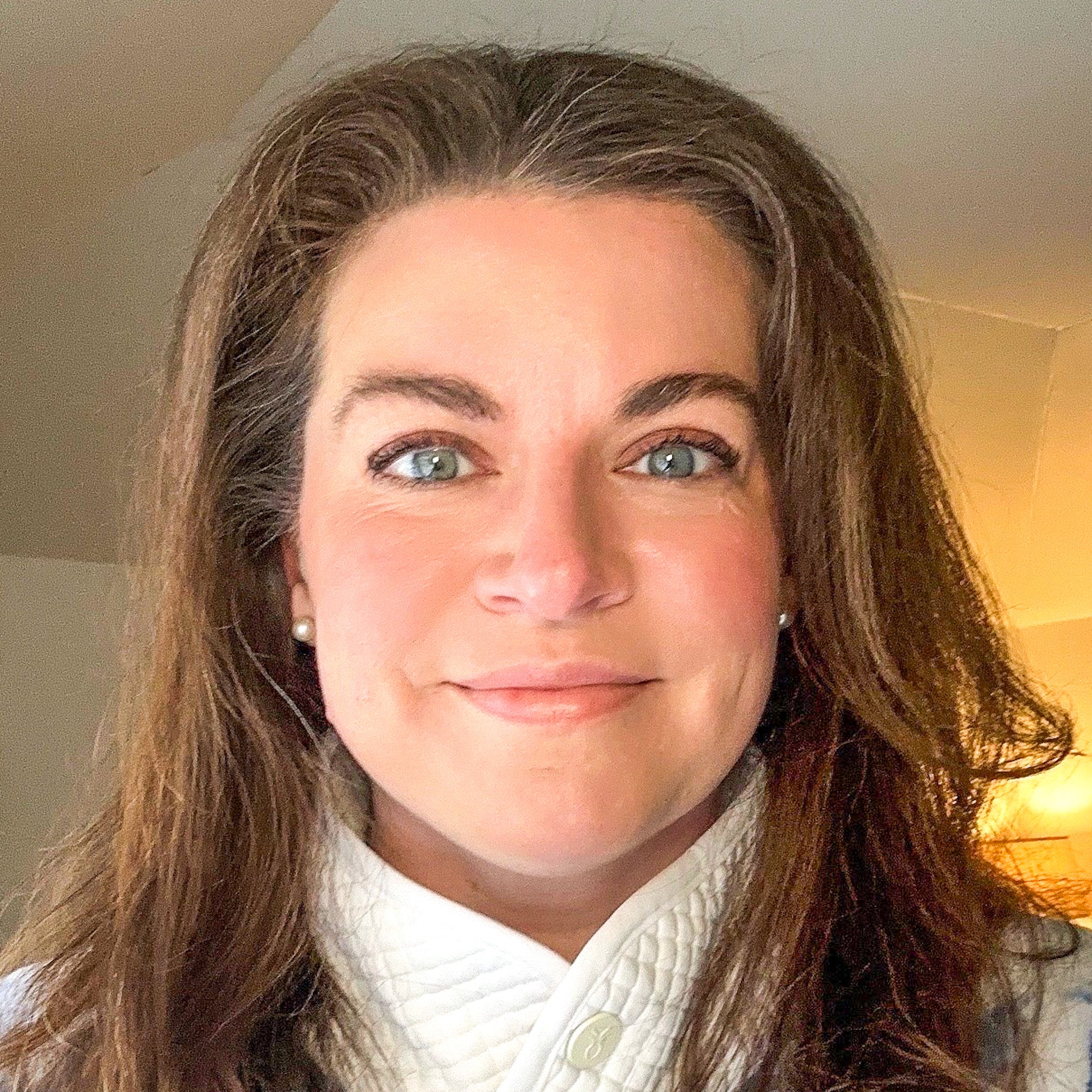 A close-up portrait of a woman with long brown hair, blue eyes, and a small smile, wearing a white textured top. The background is a softly lit indoor room with warm light.
