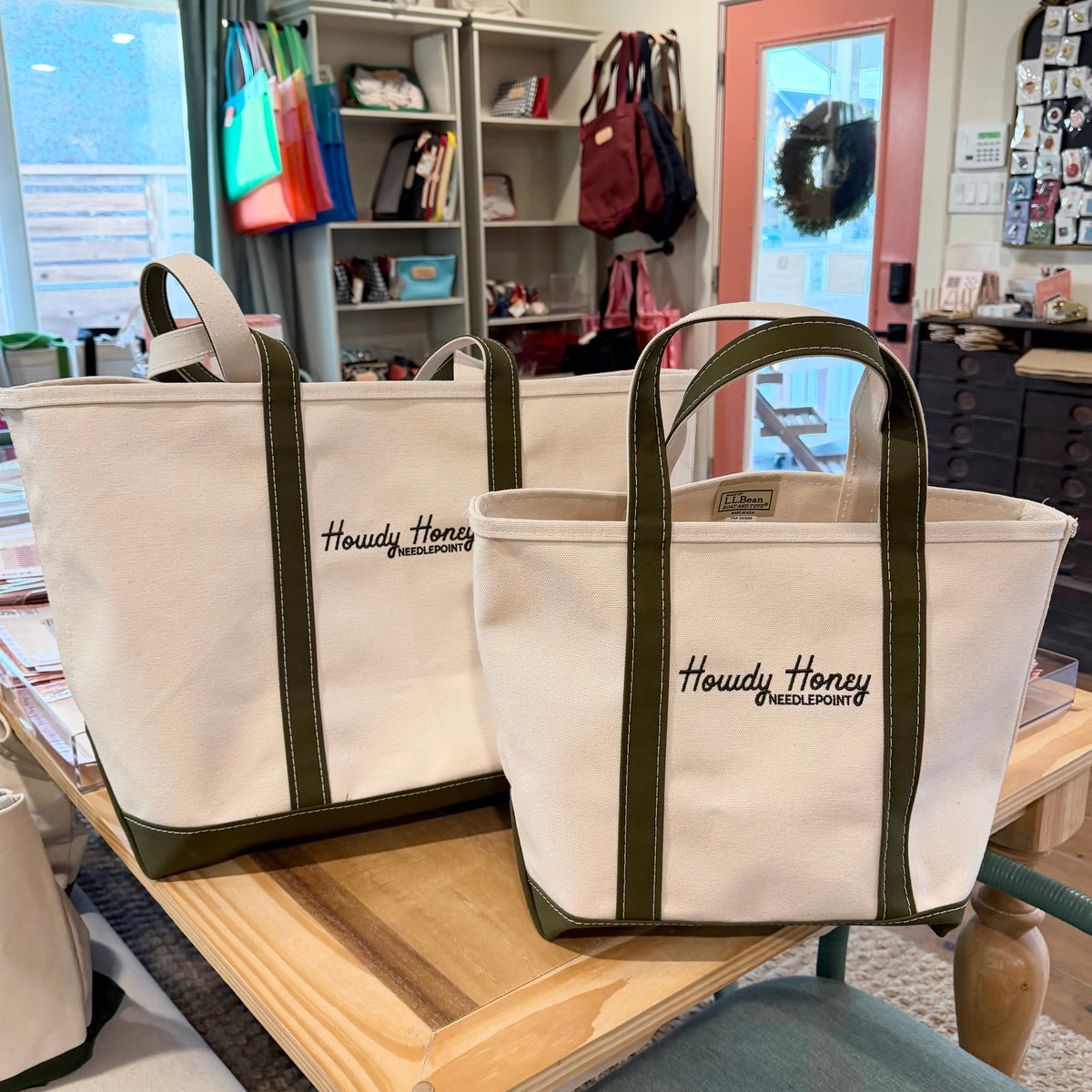 Two cream canvas tote bags with olive green trim and handles, featuring the embroidered text 'Howdy Honey @mississippi', displayed on a wooden table inside a retail store.