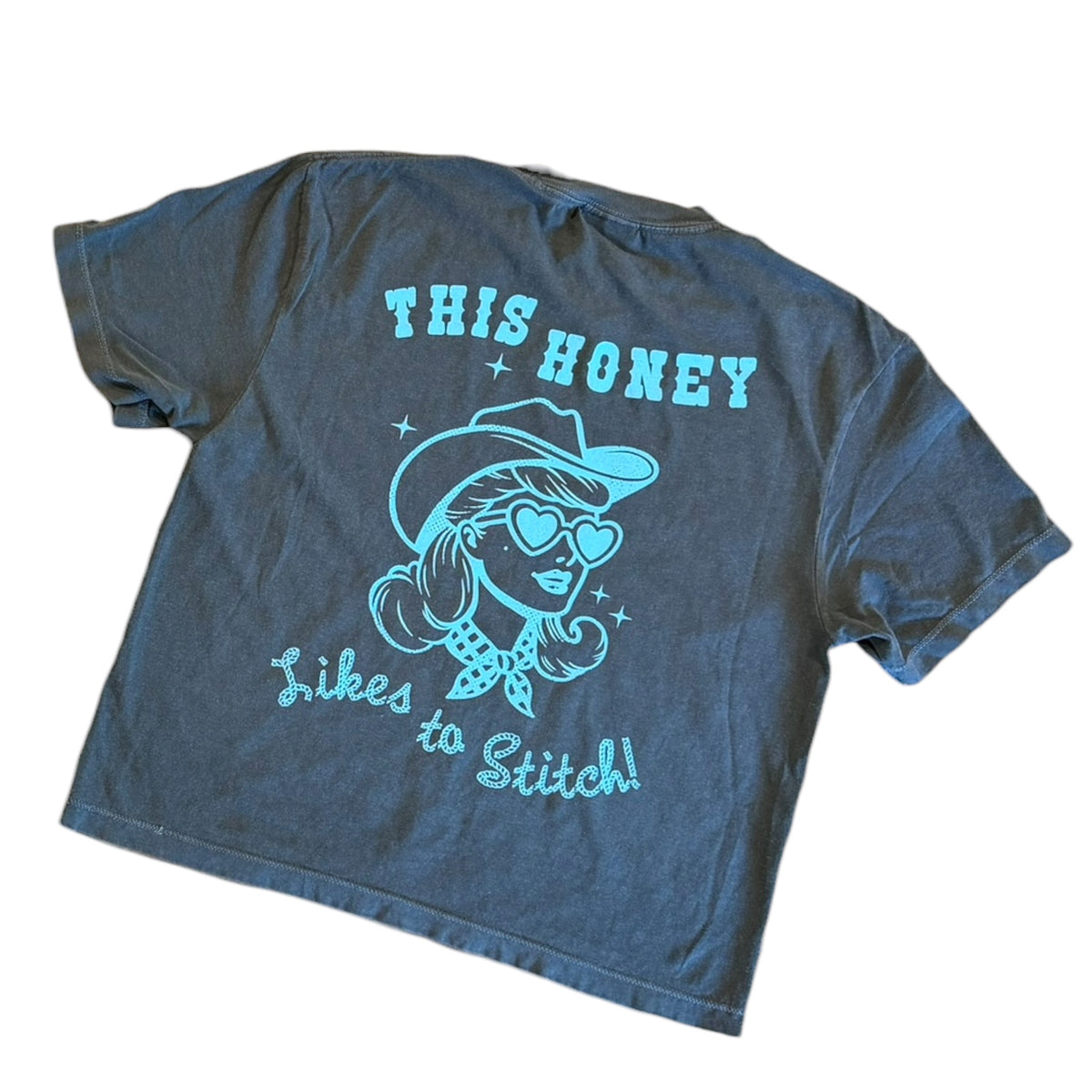 A dark gray acid-wash t-shirt with a light blue graphic on the back. The graphic features a retro-style illustration of a woman wearing a cowboy hat, heart-shaped sunglasses, and a bandana, with the text 'THIS HONEY likes to Stitch!' written in light blue.