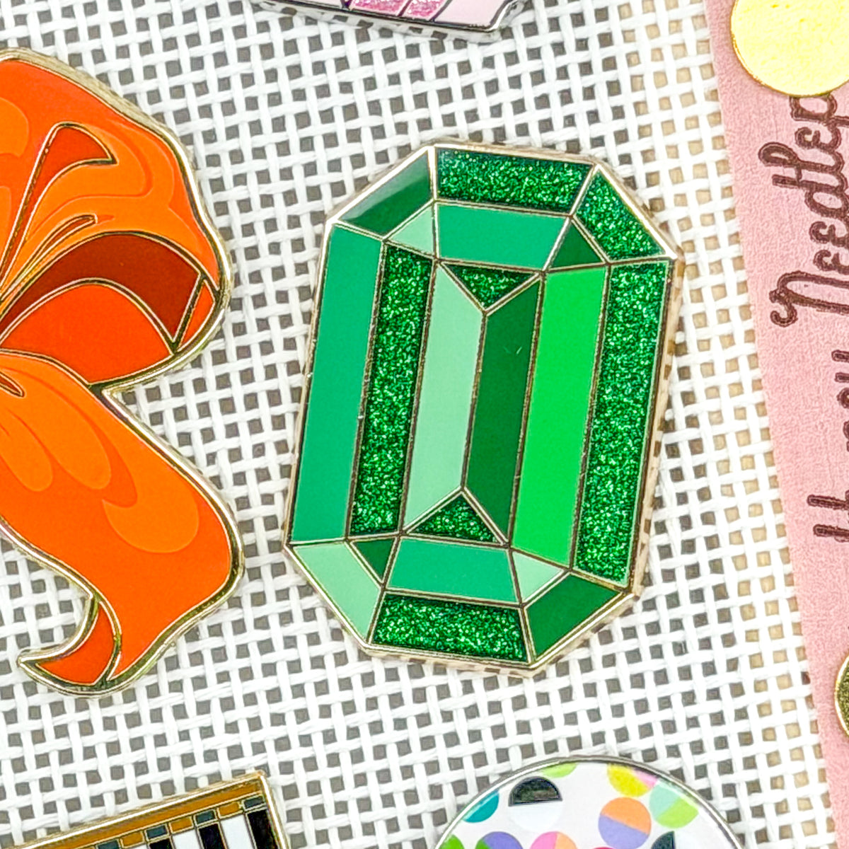 A green faceted gemstone enamel pin with gold-toned metal outlines, displayed on a white grid-patterned background alongside other colorful pins.