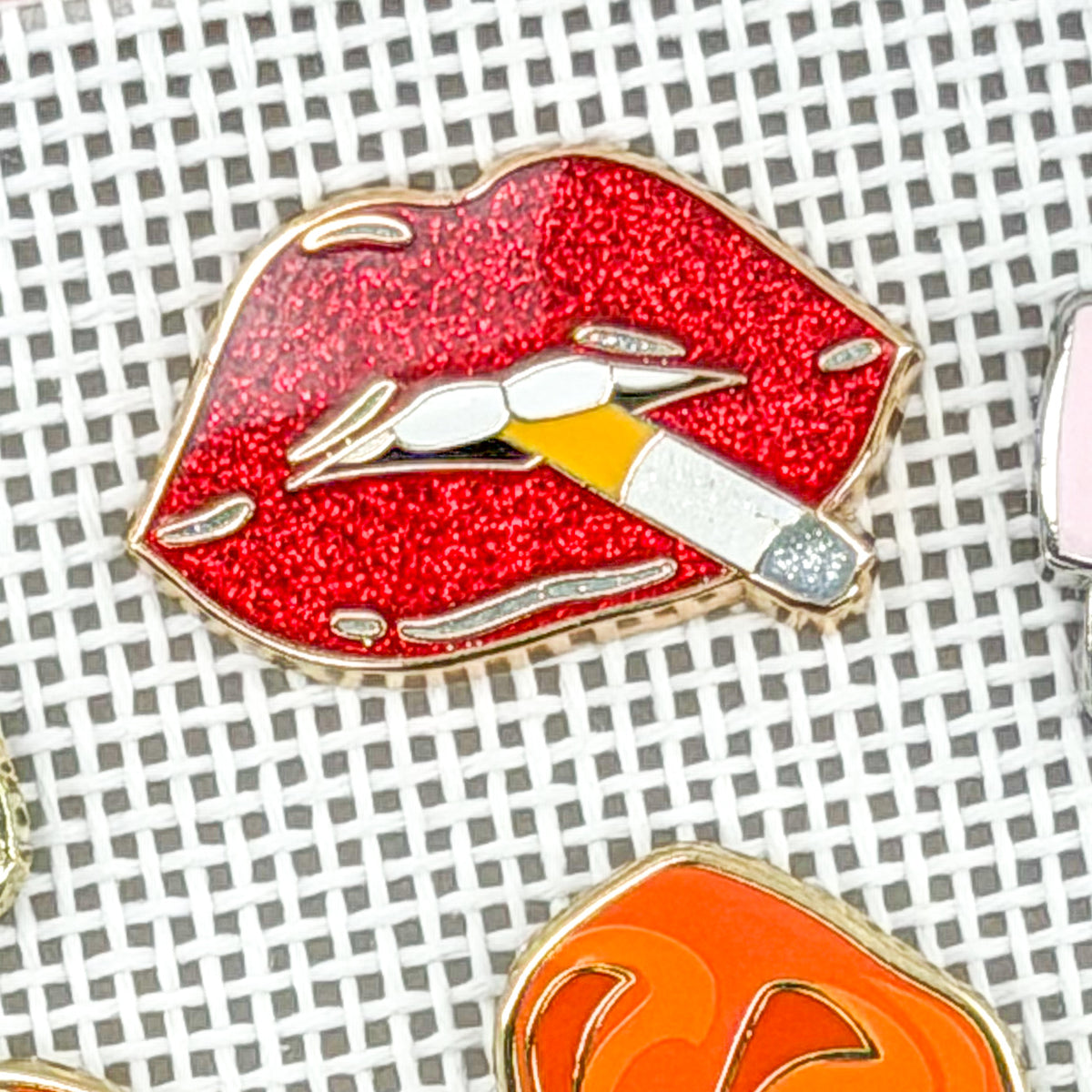 A close-up of an enamel pin shaped like red glitter lips with a silver cigarette in its mouth, placed on a white grid background.
