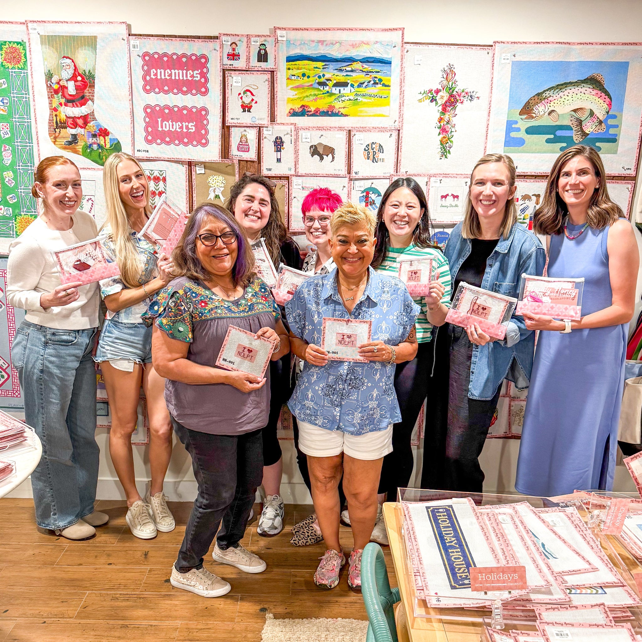 Group of ladies in front of canvas wall holding needlepoint kits.