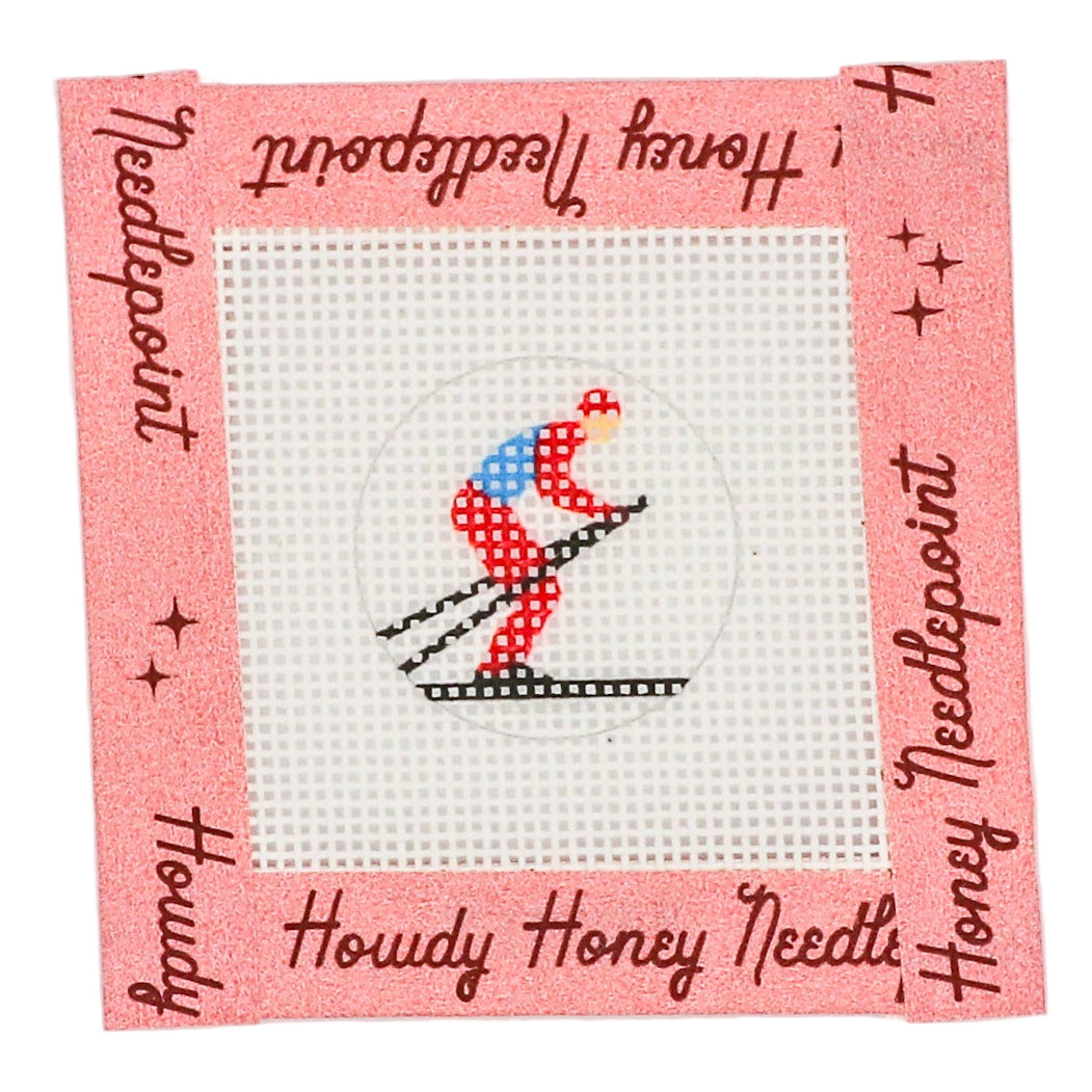 A needlecraft kit depicting a skier on a square canvas with a pink border and white background, featuring a tapestry needle and mesh fabric.