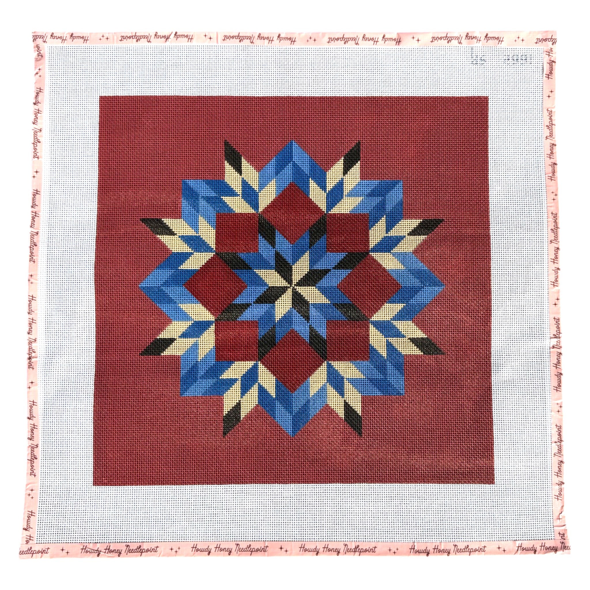 A square needlepoint canvas featuring a traditional geometric star pattern in blue, red, cream, and black on a red background, surrounded by a white mesh border with a pink printed edge.