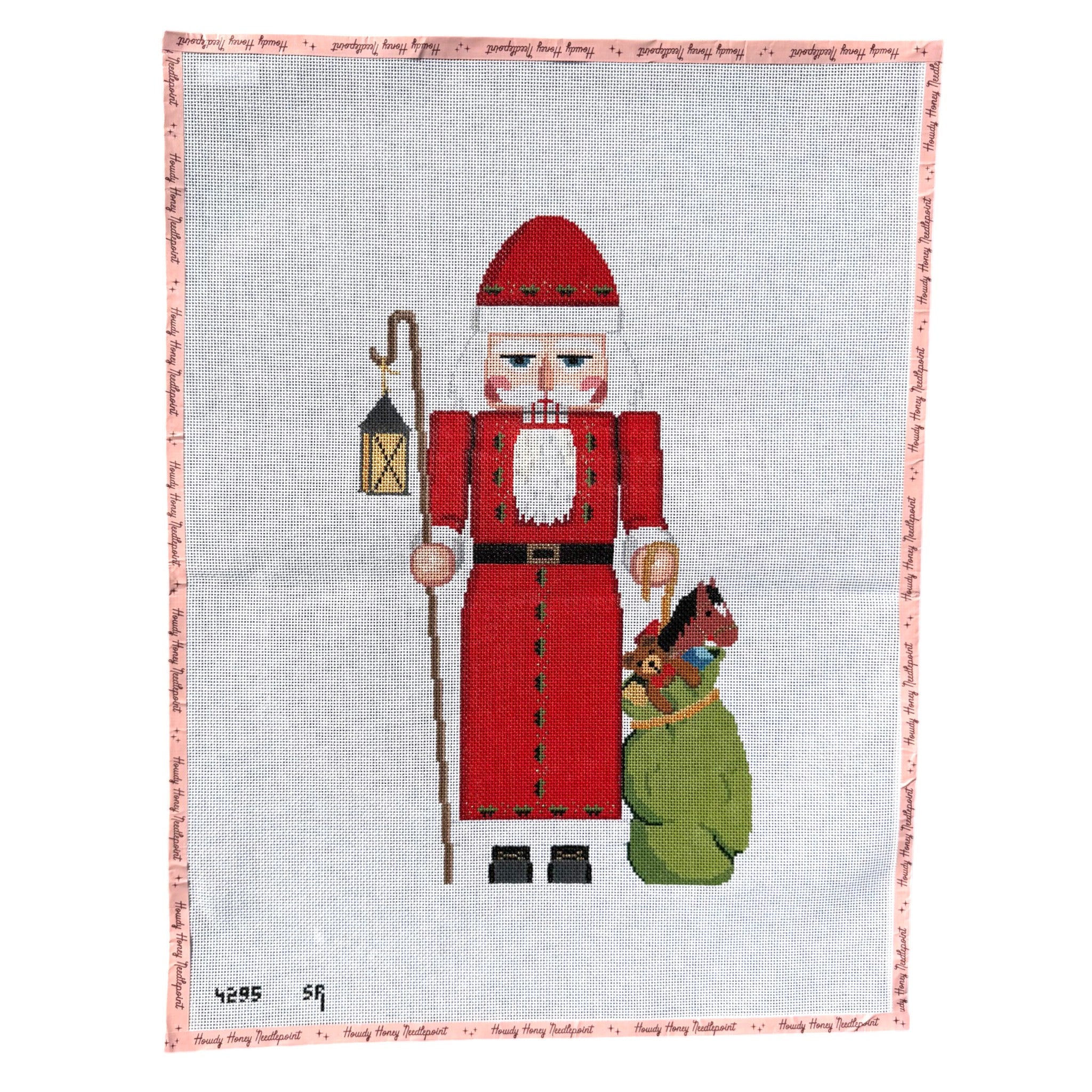 A needlepoint canvas featuring a Christmas nutcracker in a red suit holding a lantern, with a green sack on one side and a small bird on the other, set against a light gray background with a stitched pink border.