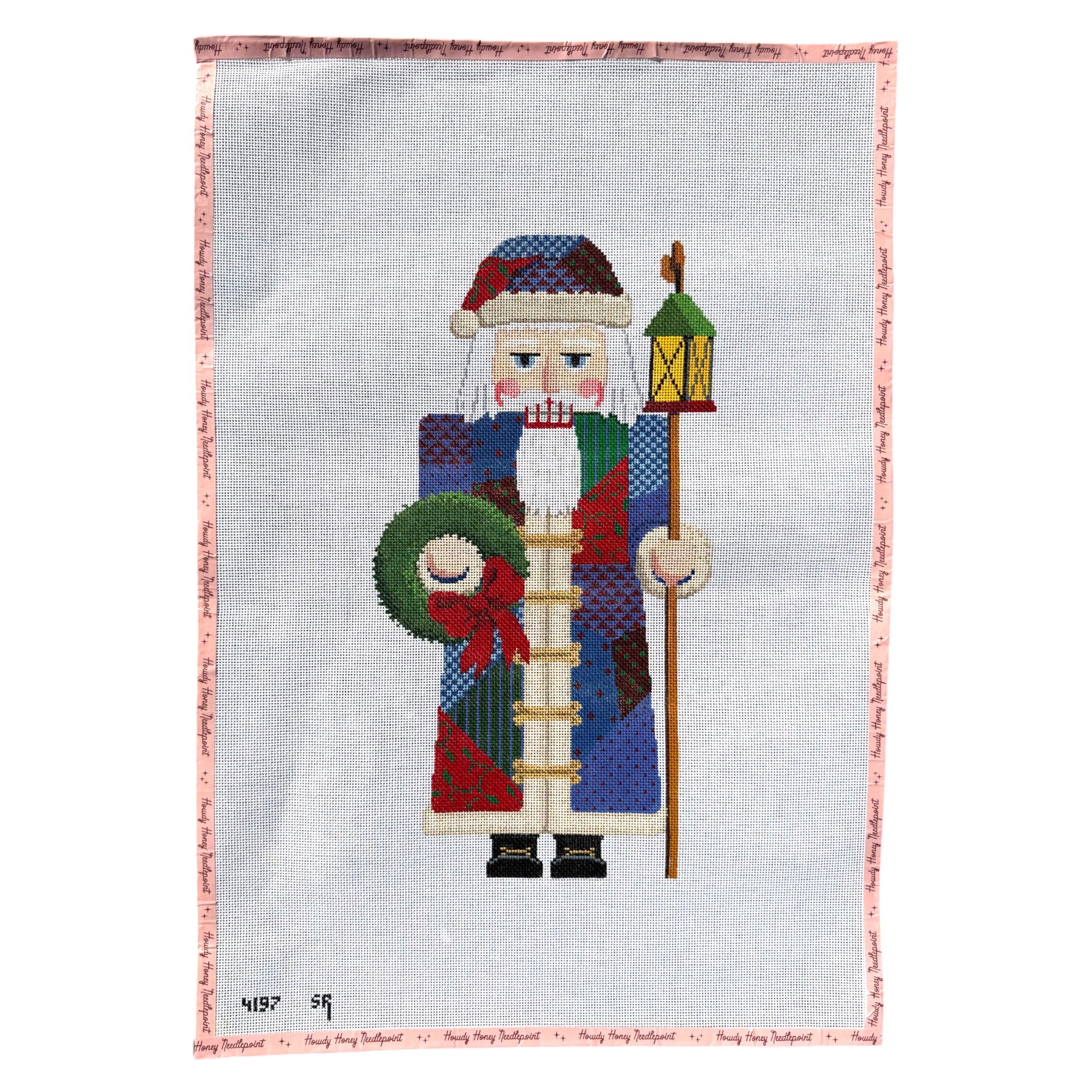 A needlepoint canvas featuring a Christmas nutcracker design. The nutcracker is dressed in a multicolored patchwork coat with blue, red, green, and beige sections, holding a green wreath and a lantern.